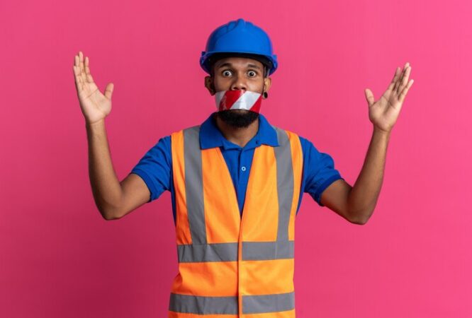 scared-young-afro-american-builder-man-uniform-with-safety-helmet-mouth-sealed-with-warning-tape-standing-with-raised-hands-isolated-pink-background-with-copy-space_141793-105559