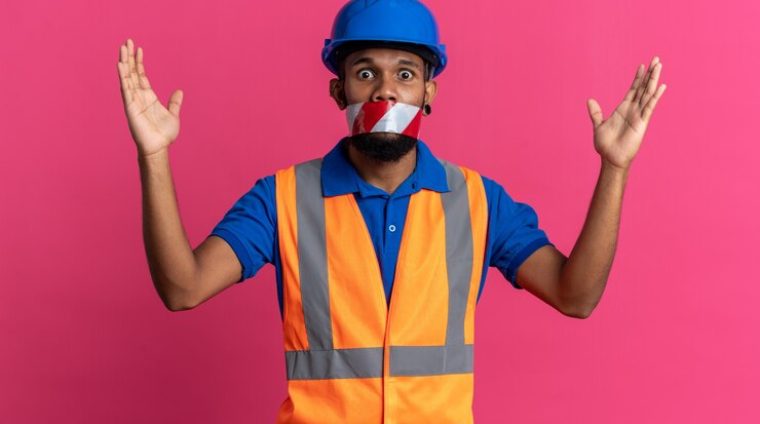 scared-young-afro-american-builder-man-uniform-with-safety-helmet-mouth-sealed-with-warning-tape-standing-with-raised-hands-isolated-pink-background-with-copy-space_141793-105559